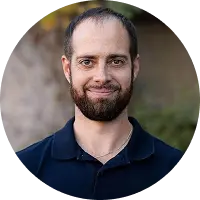 Portrait of a smiling man with a dark beard wearing a navy polo shirt outdoors circular crop