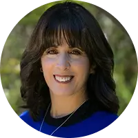 Portrait of a smiling woman with dark hair and bangs wearing a blue top outdoors with greenery in the background