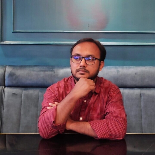 Man with glasses in a red checkered shirt sits at a dark table resting his chin on his hand in a blue booth