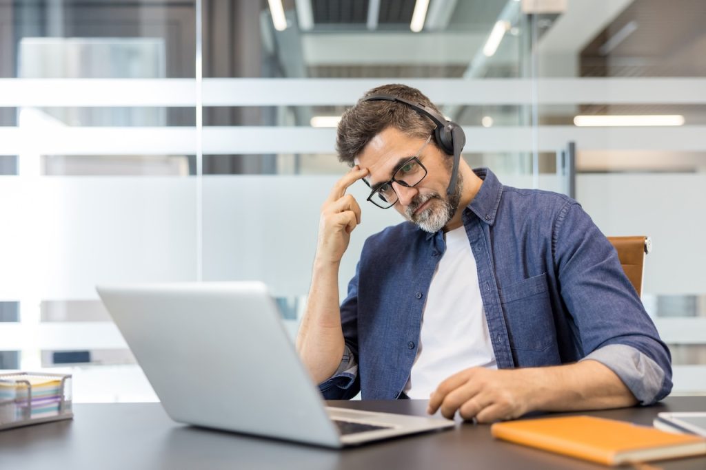 Stressed businessman wearing headset working on laptop in office trying to figure out the stats on his website without an seo audit