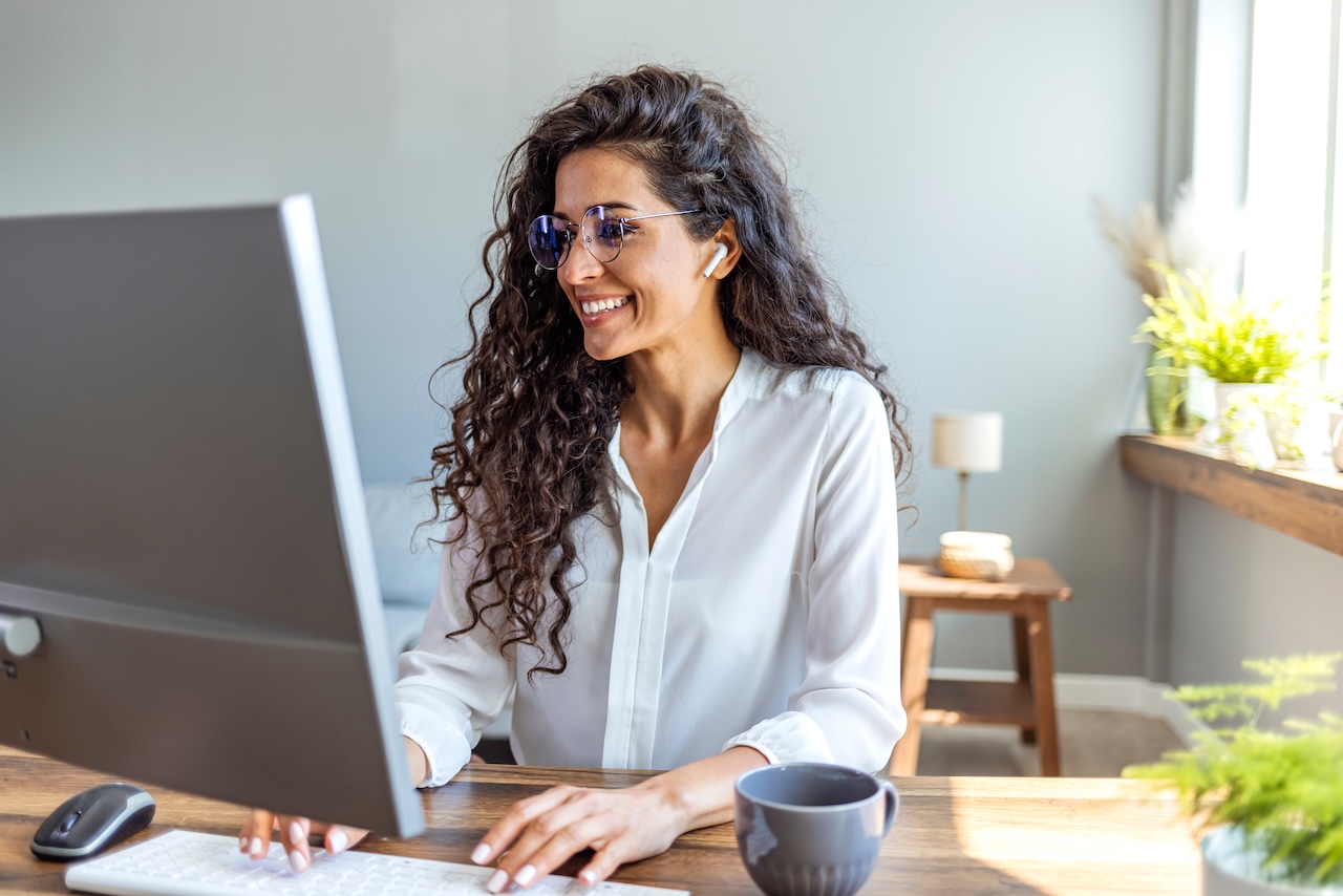 | crownsville media - seo, ai visibility & digital marketing for small business owners Woman with curly hair wearing a white blouse works at a desktop computer smiling while using wireless earbuds in a bright home office