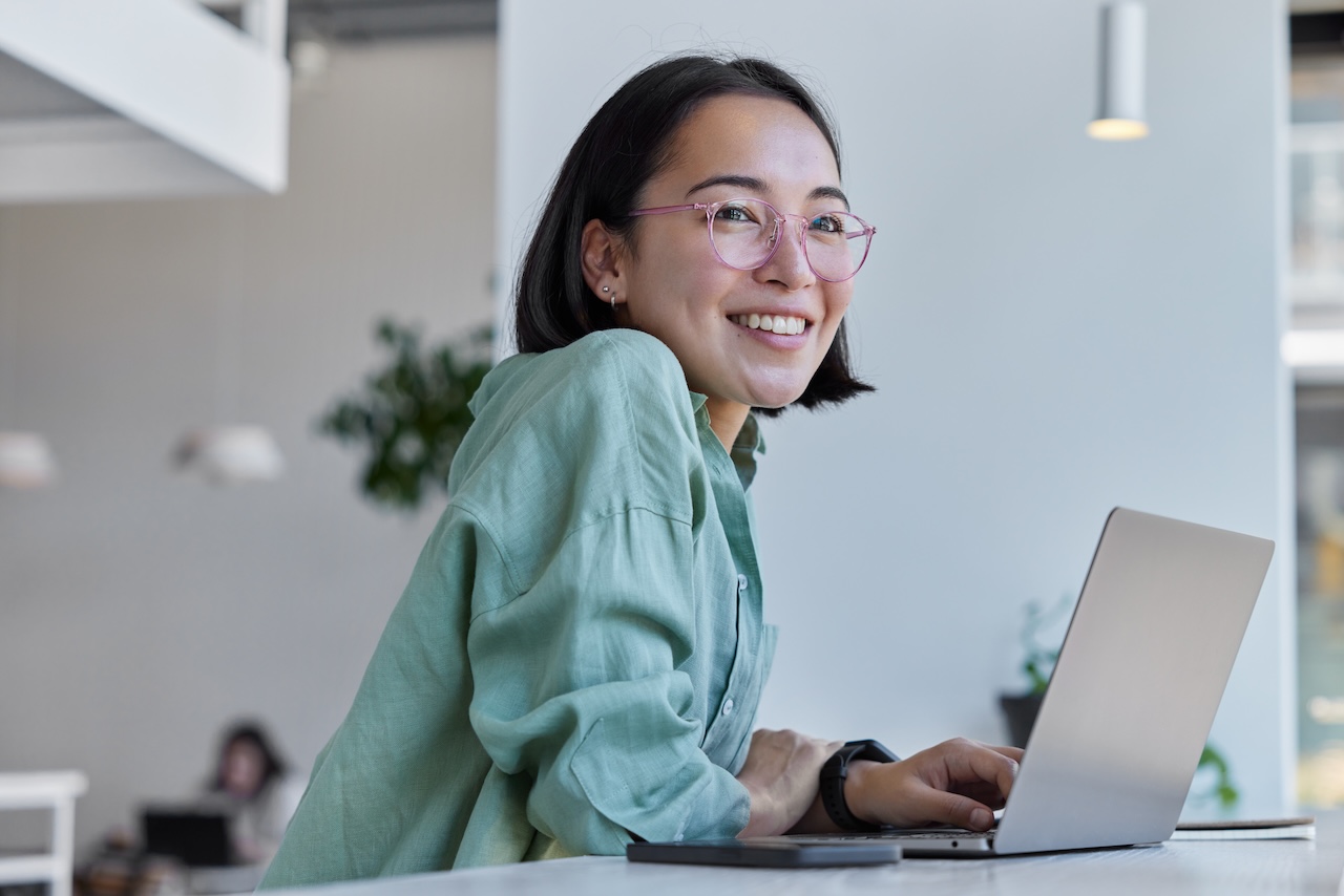 | crownsville media - seo, ai visibility & digital marketing for small business owners Smiling woman with pink glasses leaning on a laptop at a bright modern office
